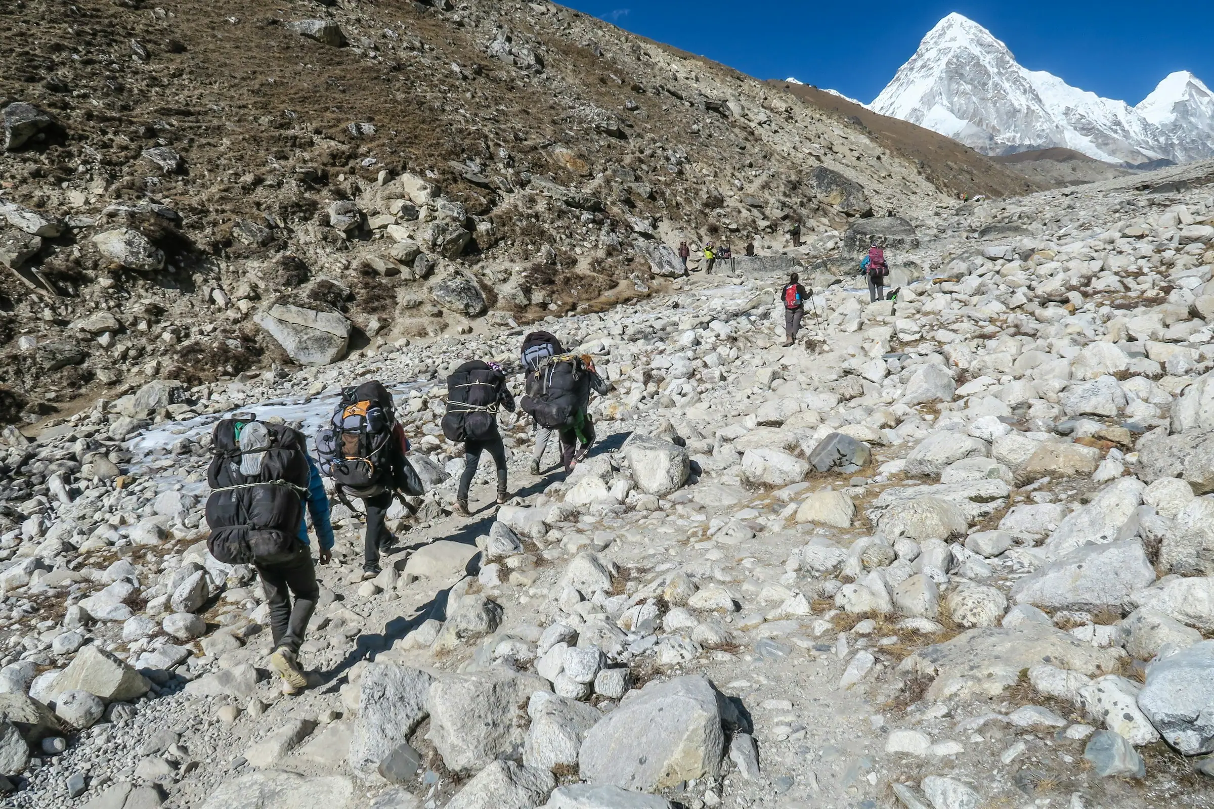 Trekkers with large packs walking up a rocky moraine trail towards a snow peak.