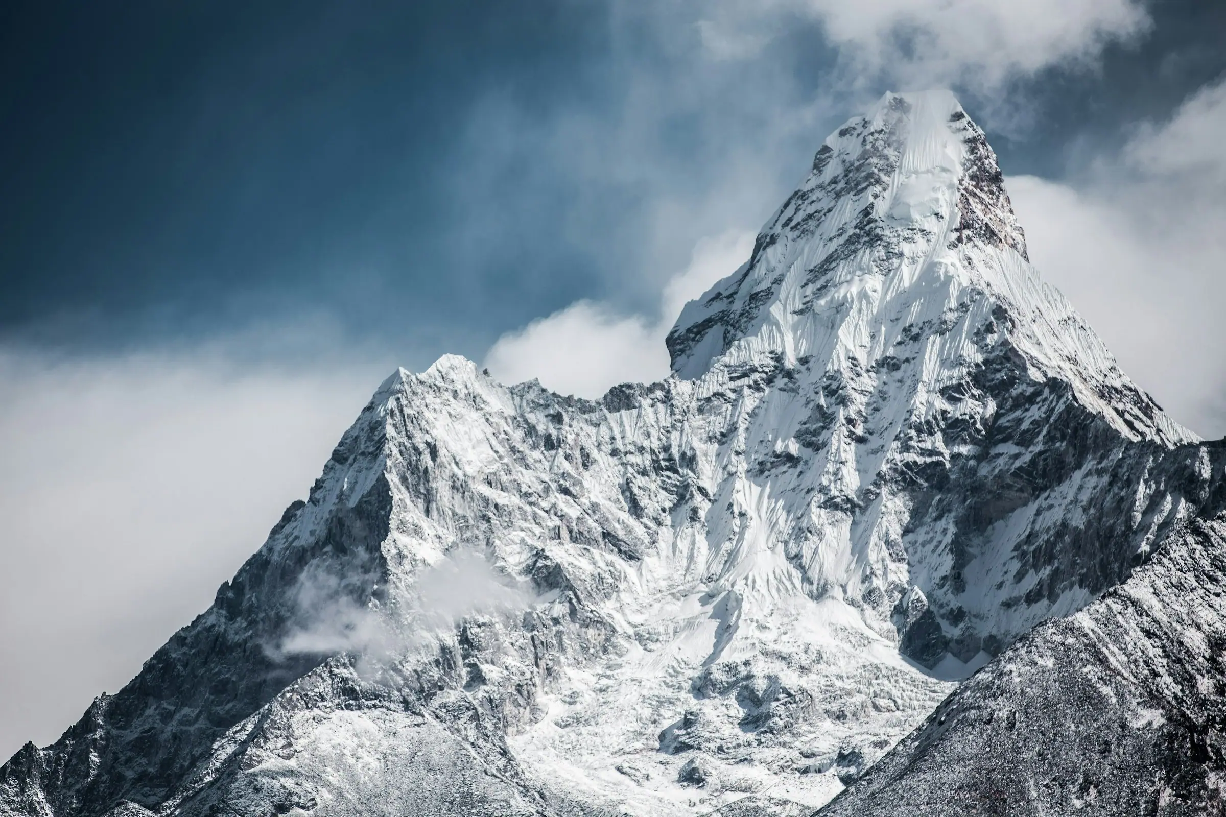 Close view of the snow-covered summit of Ama Dablam against a deep blue sky with drifting cloud on its flanks.