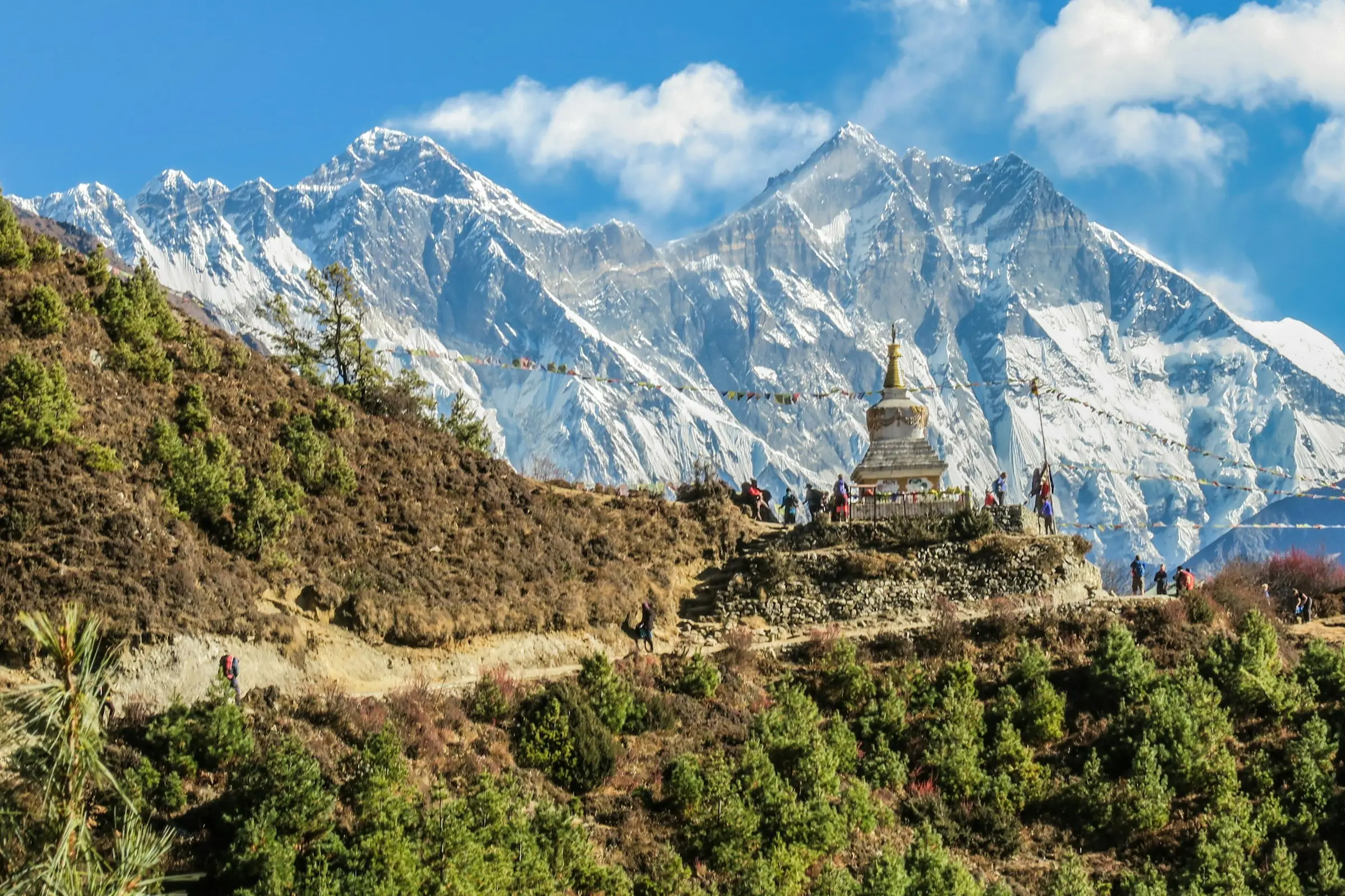 A white stupa with prayer flags on a ridge above the trail, with snow peaks behind.